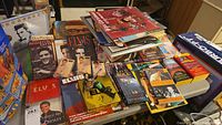 Photo of a large pile of Elvis Presley and rock music books, magazines, cassette tapes, boxed CD sets, and other collectible items on a table.