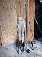 Overall group of fishing rods, some with reels, leaning against a wooden wall in a garage setting.