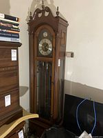 Front view of full grandfather clock showing wooden case, arched top, glass door, pendulum and weights, clock dial with Roman numerals