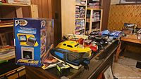 Photo showing Tyco Airblade box and Air Hogs radio control boat in yellow and black, along with vintage style RC vehicles, displayed on a surface with background shelves and storage.