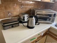 Four small kitchen appliances on countertop: Krups toaster oven, Sunbeam electric kettle, Charkescraft toaster, Heritage electric grill