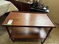 Full view of the Danish teak bar cart with two shelves on wheels. The wood grain and finish are visible. No legs appear detached in this photo but wear is noted.