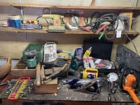 Shelves with assorted tools including wrench set, clamps, sander, hedge trimmer, jumper cables, wire wheel and other assorted hand tools scattered on shelves and workbench.