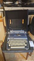 Blue-gray mechanical typewriter in dark gray hard carrying case resting on wooden stool, showing general view of lot.