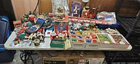 Wide view of table filled with various Christmas decorations, boxed ornaments, figurines, wrapping paper, mugs, candles, and bags.
