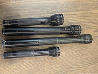 Four black aluminum flashlights laid out on wooden surface with visible wear and textured grips. Varying length, larger heads with clear lenses.
