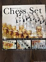 Photo of the original packaging showing the set assembled with amber and clear liquid in shot glasses representing chess pieces. Clear shot glasses have black chess piece images.