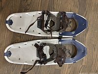 Top view of two lightweight aluminum snowshoes with white frames, blue tips, and brown nylon adjustable straps, showing bindings and underside cleats.