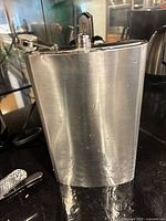 Large stainless steel flask with screw-on attached cap, curved rectangular shape, brushed steel finish, few small dents.