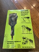 Front of the green Dremel electric engraver box showing engraver tool visible through window, usage illustrations, and list of materials it can engrave on.