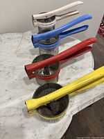 Four vintage potato ricers in white, blue, red, and yellow handles aligned on a marble surface