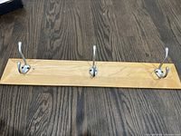 Wooden wall rack with three metal hooks shown from front angle on the floor with dark wood grain background