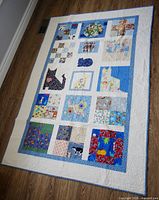Full view of quilt laid flat showing patchwork of cat and floral designs with beige and blue bordered edges.