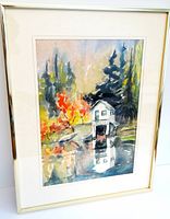 Framed watercolour artwork of a boathouse scene with autumn trees and reflection in water.