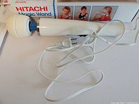 Front angle of Hitachi Magic Wand model HV-25OR massager showing the rounded vibrating head, handle, and attached power cord, with original box in background.