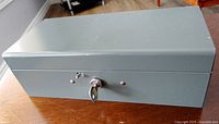Closed grey metal cash box with key inserted in lock, showing the overall exterior condition and locking mechanism.