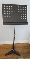 Front view of black metal perforated music stand with tripod base on wood floor against light wall.