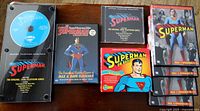 Photo shows entire Superman collection including a blue CD in a display case titled Adventures of Superman, a jewel case CD with similar title, a red cassette box labeled Superman on Radio with 4-cassette set (1 tape missing), The Complete Superman Collection DVD and 4 DVDs of Superman The Theatrical Serials Collection.