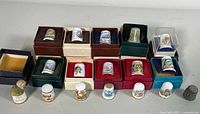 Display of 16 vintage thimbles. Fifteen porcelain thimbles mostly housed in individual display boxes, and one embossed metal thimble visible on the right side. Boxes vary in color including red, green, blue, beige, and dark hues.