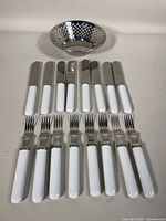 Complete set of 8 forks and 8 knives with white handles laid flat in two rows, bowl positioned behind them. Clear view of flatware style and handle color.