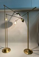 Pair of modernist brass floor lamps with adjustable arms and shades, lamps lit showing working bulbs.