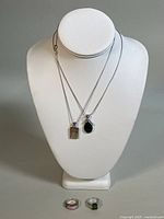 Two silver chain necklaces displayed on mannequin bust with two rings placed below. Pendants and ring stones visible.