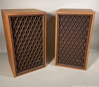 Pair of Realistic Nova-7B speakers side by side showing wood veneer finish and diamond-pattern brown grille with Realistic logo badges.