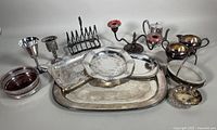 Photo of assembled vintage silver plated servingware including trays, toast rack, candlesticks, cream and sugar set, and small bowls.