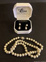 Pearl necklace coiled on black surface with matching pearl drop earrings in white Lotus Classic box showing brand and interior lining.