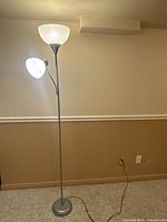 Full view of the tall floor lamp with two white shades, one torchiere and one adjustable side light, gray metal shaft, standing on carpeted floor near a beige wall.