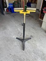 Full view of adjustable saw stand from a side angle showing yellow roller section, metal height adjustment pole with foam grip, and black tripod base.
