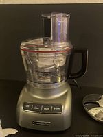 Silver KitchenAid food processor base with clear bowl and lid on black surface, showing control buttons and handle.