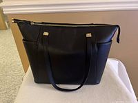 Front view of large black handbag with gold hardware on shoulder strap attachments, showing smooth faux leather material and slight wear.