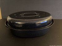 Large black enamel-coated roasting pan with dome lid standing upright, showing speckled finish and lid handle.