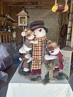 Front view of the large snowman decoration with three bear children dressed in plaid and checkered winter clothing, standing on a decorated base.