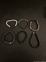 Six lava rock bead bracelets in various colors and styles on black background.