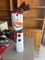 Full view of the wooden snowman log showing painted white body, black buttons, smiling face with orange carrot nose, and red-green plaid hat and scarf.