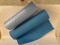 Blue yoga mat rolled out with gray foam roller placed alongside on a beige surface, showing texture and color of both items.