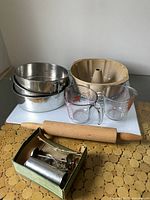 Photo showing all baking items including two metal cake pans, one glass bundt cake pan, two glass measuring cups, a wooden rolling pin, and cookie cutter set in box on white surface.