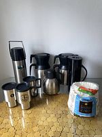 Photo showing three stainless steel coffee decanters with black lids and handles, three matching travel mugs, one small round thermos, and a pack of white coffee filter papers.
