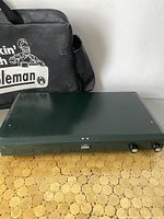 Closed green Coleman propane stove with two control knobs and a latch at front, set on a hexagonal wooden surface next to a black zippered case with Coleman logo.