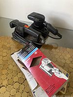 Two black electric hand sanders with power cords on circular patterned surface with some sandpaper packages in front