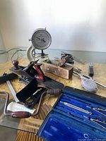 Wide shot of the vintage tool lot showing dial micrometer, hand planers, adjustable squares, hand drill, measuring tape, and drafting tools in blue case on a glass table.