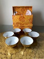 Twelve small gold plated bowls with white interior displayed in front of two original floral-patterned boxes.