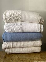 Five blankets stacked; two white blankets with waffle knit texture on one end at top, middle blue wool blend blanket, and two cream-colored chunky knit blankets at bottom.