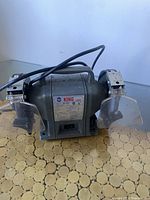 Front view showing the full bench grinder with both grinding wheels and safety guards visible, placed on a wood-patterned surface.