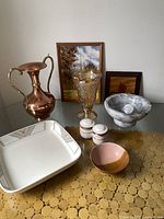 Full lot on table showing all items including copper vase, stone mortar and pestle, golden lustre vase, ceramic bowl, two small jars, two framed artworks, and serving dish