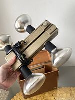 Top angle view of the vintage 4-light lighting bar showing metal reflectors, gold-tone metal body, and attached power cord. The owner's hand holds the bar above the carrying case.