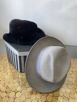 Women's dark mink and sable fur hat placed on top of a striped original box, alongside a men's light beige felt hat with wide gray ribbon band.