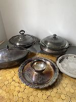Group photo of three silver plated covered serving dishes, one ornate large round tray with central cup, and another silver plated round tray, all displayed on a yellow textured surface.